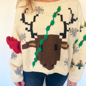 May include: A white knitted sweater with a brown reindeer design. The reindeer has green holly leaves around its antlers. The sweater has a red bow on one sleeve.