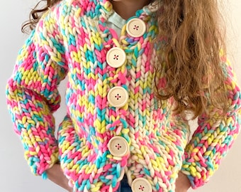 May include: A chunky knit cardigan in a vibrant mix of pink, yellow, blue, and white. The sweater features large wooden buttons and a hooded design. The colorful, textured knitwear is a cozy and stylish garment for children.