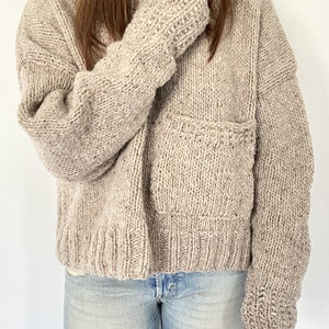 May include: A beige knitted cardigan with a patch pocket and long sleeves. The cardigan is made from chunky yarn and has a relaxed fit.