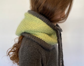 May include: A knitted hood in shades of green and gray. The hood is a light green color on the inside and a darker gray on the outside, with a gray trim. The hood has a drawstring and is worn over a dark gray coat.