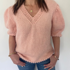 以下が含まれることがあります： Vネックと短いパフスリーブが付いた薄いピンクのニットセーター