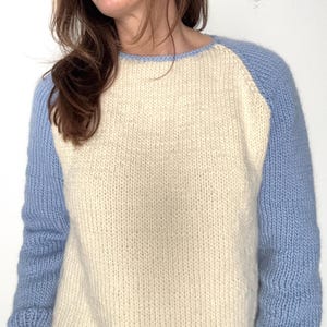 May include: Cream-colored knit sweater with light blue raglan sleeves. The sweater has a crew neck and a textured knit pattern. The sleeves and neckline are a solid light blue, contrasting with the cream body. A cozy, casual top.