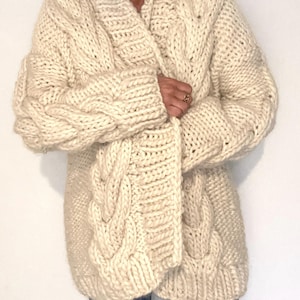 May include: A cream-coloured, chunky knit cardigan with a shawl collar and cable knit detailing. The oversized sweater has long sleeves and a relaxed fit. A cosy, warm, and stylish outerwear garment for cooler weather.