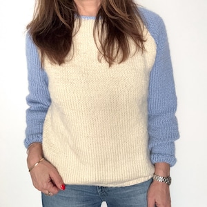 May include: Cream-colored sweater with light blue raglan sleeves. The knitwear features a crew neck and ribbed cuffs. The sweater is made of a soft, textured material and is paired with blue jeans.