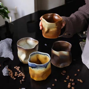 Könnte beinhalten: Vier handgefertigte Keramikbecher mit einzigartigen, organischen Formen und Erdtönen. Die Becher weisen eine Mischung aus braunen, beigen und blauen Glasuren mit einer strukturierten, unebenen Oberfläche auf. Kaffeebohnen sind verstreut.
