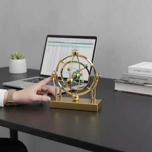 May include: A gold-colored desk ornament with a spherical design featuring green and gold spheres and rings. The ornament sits on a rectangular base. A hand is touching the ornament. A laptop and books are in the background.