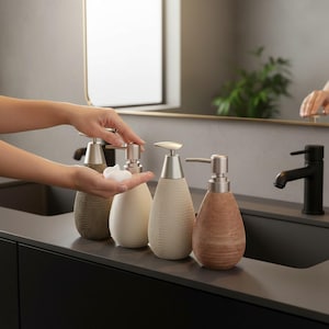 Puede incluir: Cuatro dispensadores de jabón líquido decorativos con exteriores texturizados, similares a tejidos, en tonos beige, marrón y bronceado. Cada dispensador tiene una bomba de color plateado. Se está utilizando un dispensador para dispensar jabón.