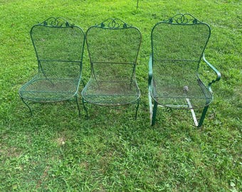 May include: Three green metal garden chairs with a decorative back design and mesh seating. The chairs are placed on a grassy lawn. One chair has armrests.