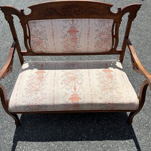 May include: A vintage wooden love seat with a floral patterned fabric upholstery. The seat has a carved wooden frame and scrolled arms.