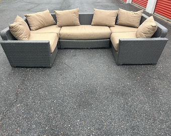May include: A U-shaped outdoor sectional sofa with a dark gray woven frame and tan cushions. The sofa includes several matching tan throw pillows. The furniture is placed on a dark asphalt surface.