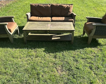 May include: Outdoor furniture set on a grassy lawn. The set includes a two-seater sofa, two armchairs, and a coffee table. The frames are light wood with woven sides and brown cushions. The coffee table has a slatted top.