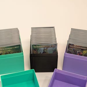 Könnte beinhalten: Eine Sammlung von Sammelkarten-Aufbewahrungsboxen in Türkis, Schwarz und Lila. Jede Box enthält einen Stapel Karten, von denen einige sichtbar sind. Die Boxen sind leer, ohne Deckel, und auf einer weißen Oberfläche angeordnet.