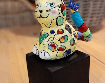 Può includere: Una stravagante statuetta a forma di gatto con un corpo giallo ornato di macchie colorate e una coda multicolore. Il gatto è appoggiato su una base quadrata nera, con un dettaglio a farfalla sulla coda. Il gatto ha una faccia sorridente.