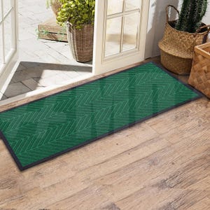May include: A green doormat with a chevron pattern and black edging. The doormat is placed on a wooden floor.