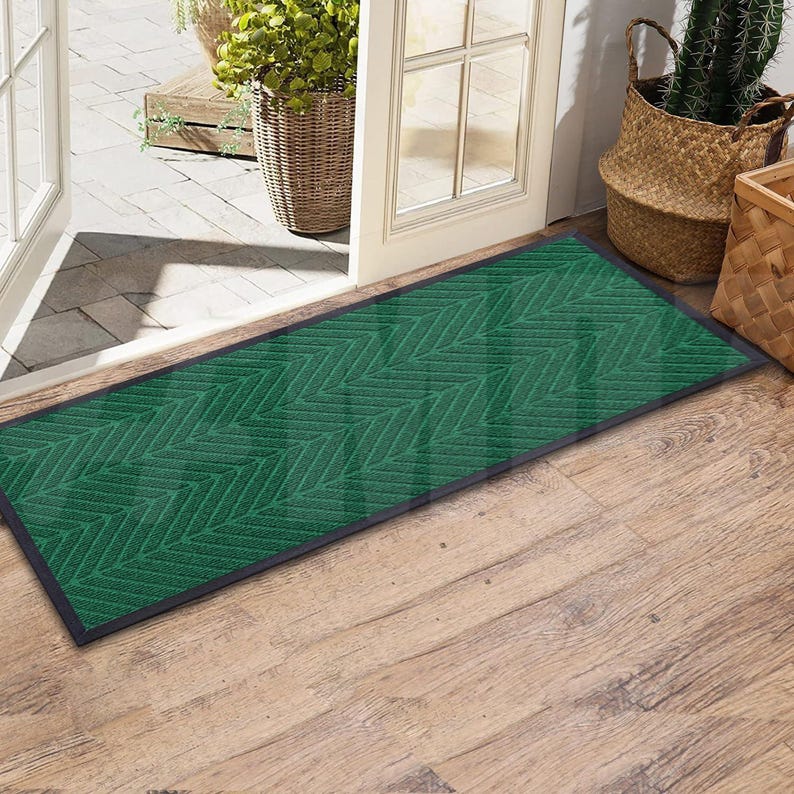 May include: A green doormat with a chevron pattern and black edging. The doormat is placed on a wooden floor.