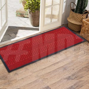 May include: A red doormat with a chevron pattern and black edging. The doormat is placed in front of a door with a wooden frame.