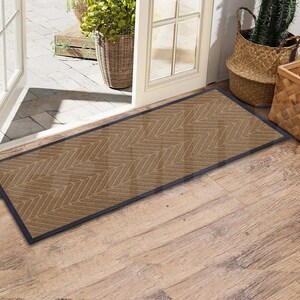 May include: A brown doormat with a herringbone pattern and a black border. The doormat is placed in front of a door with a wooden frame.