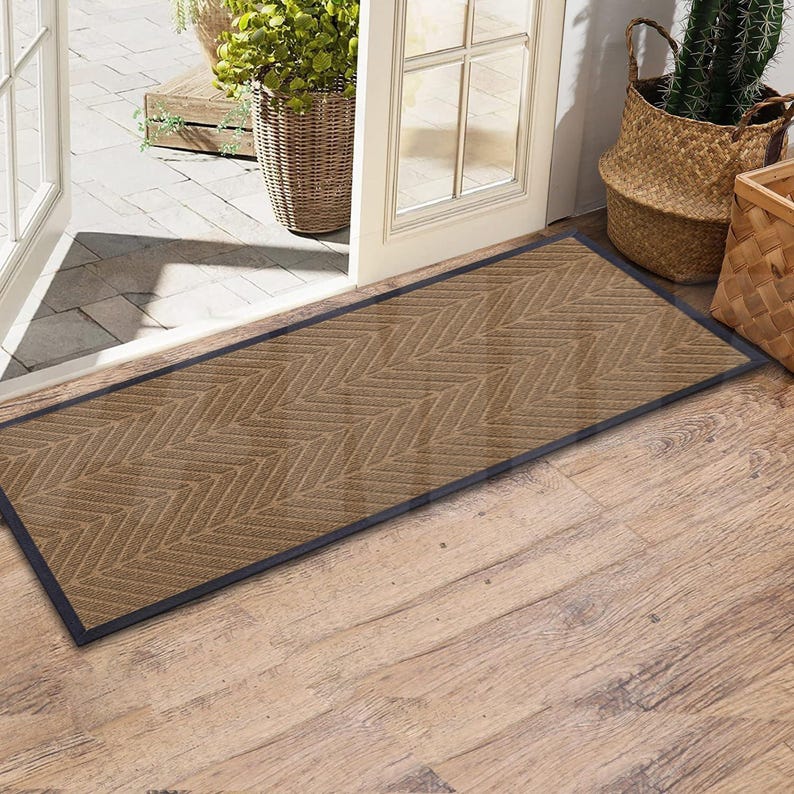 May include: A brown doormat with a herringbone pattern and a black border. The doormat is placed in front of a door with a wooden frame.