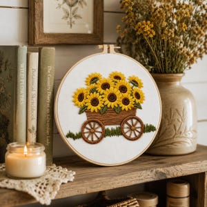 May include: A circular embroidery hoop featuring a hand-stitched design of a brown wooden cart filled with bright yellow sunflowers with dark brown centres. The cart sits on a patch of green grass, all set against a plain white fabric background.