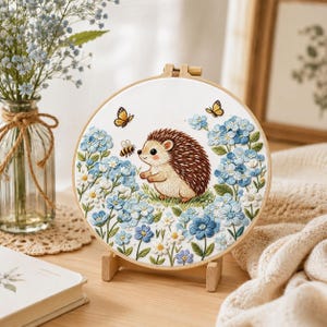 May include: A circular wooden embroidery hoop containing a white fabric panel with a hand-stitched design of a brown hedgehog surrounded by blue hydrangea flowers, white daisies, a small yellow bee, and two orange butterflies on a light background.