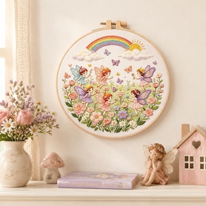 Puede incluir: Un bastidor de bordado circular que presenta una escena cosida a color de cinco hadas con alas en un jardín de flores, mariposas y un arcoíris con un sol sobre nubes blancas. El diseño está realizado sobre tela blanca y enmarcado en madera.