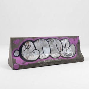 Könnte beinhalten: Betonsperre mit Graffiti-Kunst. Die graue Sperre zeigt ein großes, silbernes und schwarzes Graffiti-Design mit lila Umrandung und kleinen lila Herzformen. Das Kunstwerk ist auf der Vorderseite der Sperre zentriert.