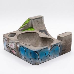 K&ouml;nnte beinhalten: Beton-Aschenbecher in Form eines Mini-Skateparks. Der graue Aschenbecher hat eine Rampe, Treppen und ein Becken mit blauen und rosa Graffiti. Die Rampe ist mit gr&uuml;nem Kunstrasen versehen.
