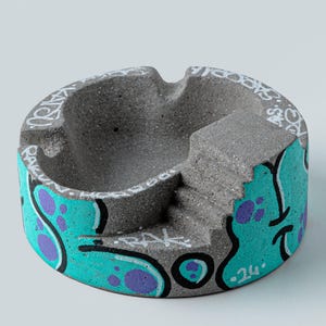 Puede incluir: Un cenicero circular de hormigón gris con una superficie texturizada y un pequeño detalle de escalera. El exterior está decorado con patrones de pintura estilo grafiti en turquesa y morado, incluyendo el texto KATSU y 24.
