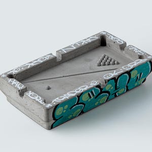May include: A rectangular grey concrete ashtray shaped like a pool table, featuring white graffiti-style text along the edges and a vibrant teal and black graphic design on the side. The surface includes a moulded triangle of small spheres.