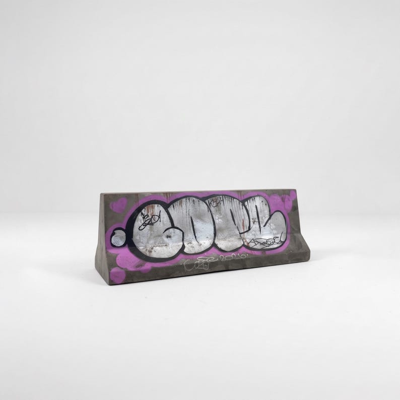 May include: Concrete barrier with graffiti art. The gray barrier features a large, silver and black graffiti design with a purple outline and small purple heart shapes. The artwork is centered on the front of the barrier.