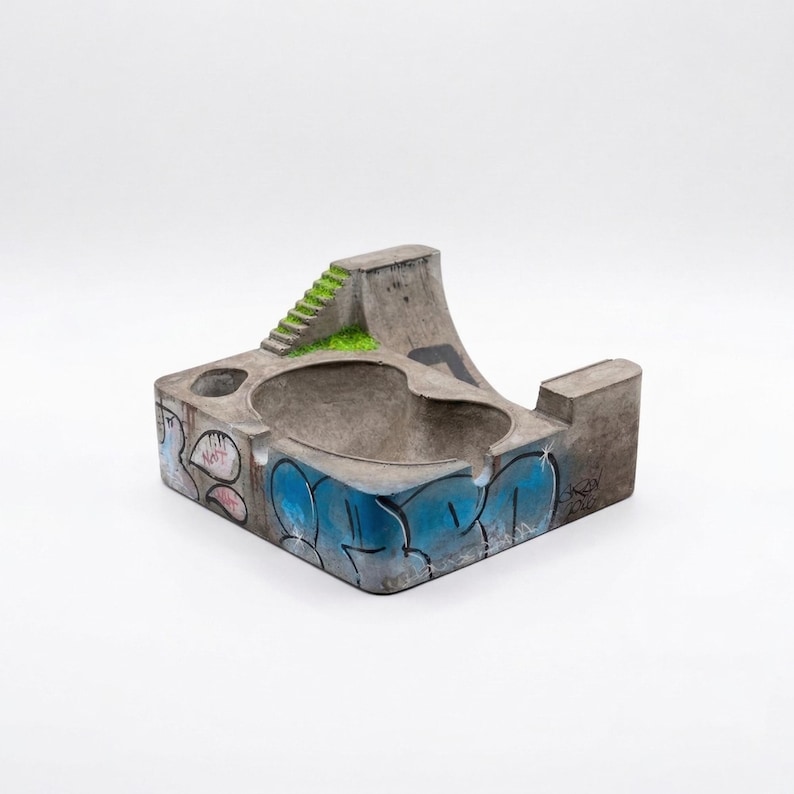 K&ouml;nnte beinhalten: Beton-Aschenbecher in Form eines Mini-Skateparks. Der graue Aschenbecher hat eine Rampe, Treppen und ein Becken mit blauen und rosa Graffiti. Die Rampe ist mit gr&uuml;nem Kunstrasen versehen.