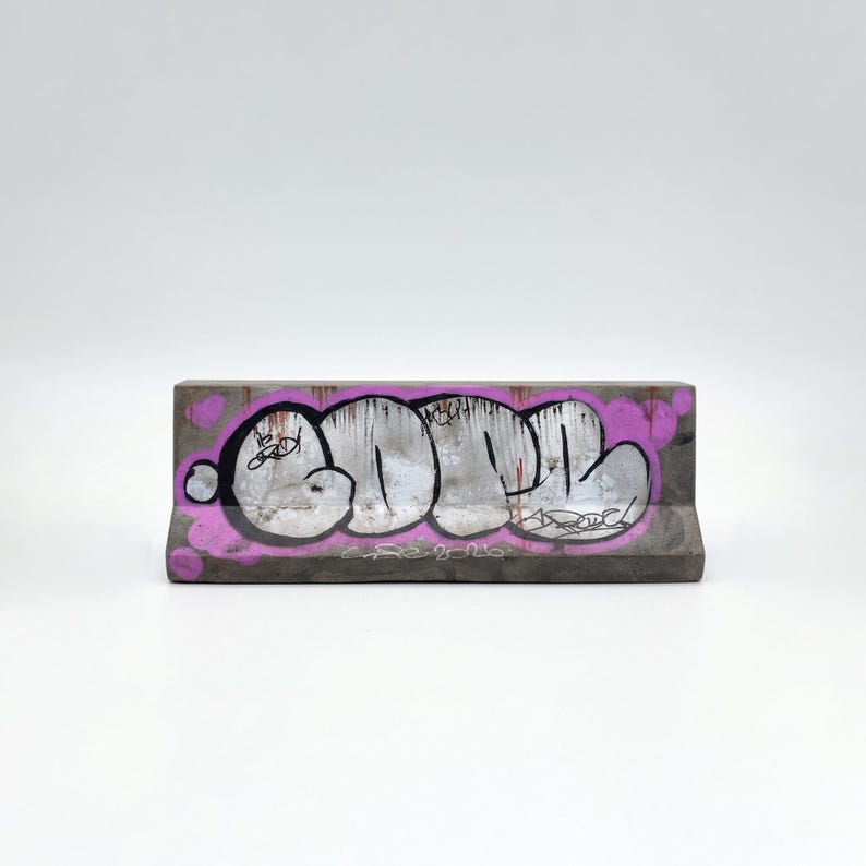 K&ouml;nnte beinhalten: Betonsperre mit Graffiti-Kunst. Das Wort "DON" ist in Wei&szlig; mit schwarzen Umrissen und einem rosa Hintergrund aufgespr&uuml;ht. Die Sperre ist grau und hat eine rechteckige Form.