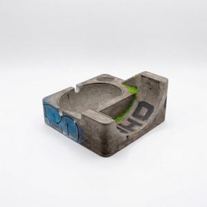 K&ouml;nnte beinhalten: Beton-Aschenbecher in Form eines Mini-Skateparks. Er verf&uuml;gt &uuml;ber eine Sch&uuml;ssel, eine Rampe und blaue Buchstaben im Graffiti-Stil. Auf der Rampe stehen die Buchstaben "WHO" in Schwarz. Gr&uuml;nes Kunstgras f&uuml;gt Details hinzu.
