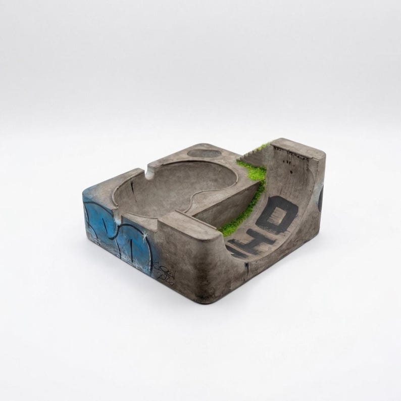 K&ouml;nnte beinhalten: Beton-Aschenbecher in Form eines Mini-Skateparks. Er verf&uuml;gt &uuml;ber eine Sch&uuml;ssel, eine Rampe und blaue Buchstaben im Graffiti-Stil. Auf der Rampe stehen die Buchstaben "WHO" in Schwarz. Gr&uuml;nes Kunstgras f&uuml;gt Details hinzu.