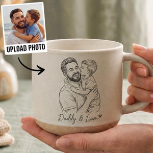 May include: Cream-colored ceramic mug with a sketch of a father and child, with the text "Daddy & Liam". The mug has a brown base and a handle. A photo with the text "UPLOAD PHOTO" is shown next to the mug.