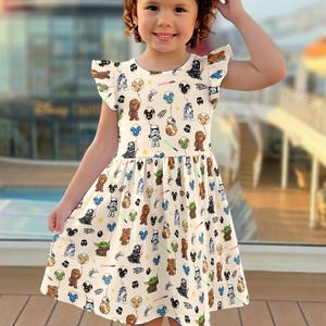 Op de afbeelding: Een cr&egrave;mekleurige jurk met een Star Wars-thema print. De jurk heeft een uitlopende rok en fladderende mouwen. De print bevat verschillende Star Wars-personages en elementen, zoals Darth Vader, Chewbacca en lichtzwaarden. De jurk wordt gedragen door een jong meisje.