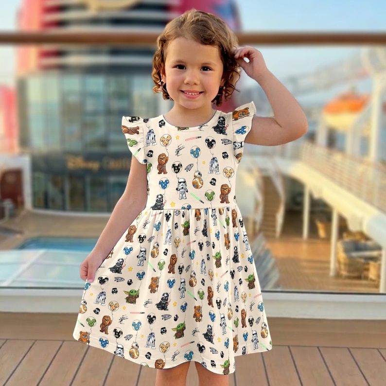 Op de afbeelding: Een cr&egrave;mekleurige jurk met een Star Wars-thema print. De jurk heeft een uitlopende rok en fladderende mouwen. De print bevat verschillende Star Wars-personages en elementen, zoals Darth Vader, Chewbacca en lichtzwaarden. De jurk wordt gedragen door een jong meisje.