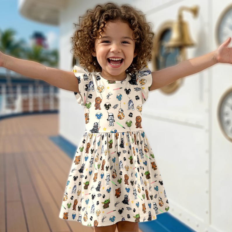 Op de afbeelding: Een cr&egrave;mekleurige jurk met Star Wars-personages en Mickey Mouse-ballonnen. De jurk heeft korte mouwen met gerimpelde randen en een uitlopende rok. De print bevat personages als Darth Vader, Chewbacca en Yoda. De jurk wordt gedragen door een jong kind.