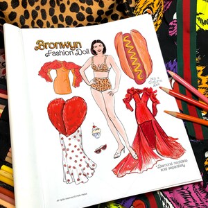 May include: A paper doll book titled "Bronwyn Fashion Doll" featuring a fashion doll with various outfits, including a leopard print bikini, a red dress, and a hot dog costume. The book is open, with colored pencils and patterned fabric in the background.