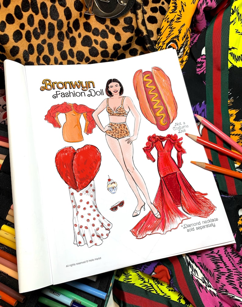 May include: A paper doll book titled "Bronwyn Fashion Doll" featuring a fashion doll with various outfits, including a leopard print bikini, a red dress, and a hot dog costume. The book is open, with colored pencils and patterned fabric in the background.
