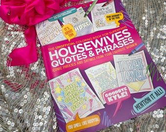 May include: A coloring book titled "Housewives Quotes & Phrases" with iconic sayings, featuring illustrations and text. A bright pink bow sits above the book on a sequined surface. The book includes phrases like "Who Gon' Check Me, Boo?" and "Mention It All!"