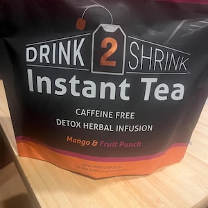 May include: Black bag of "Drink 2 Shrink Instant Tea" with orange and purple accents. The bag features the text "Caffeine Free Detox Herbal Infusion" and "Mango & Fruit Punch". The bag contains 28 stick packs.