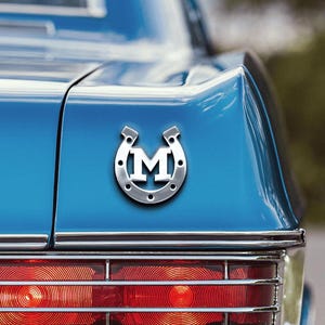 Könnte beinhalten: Nahaufnahme des Hecks eines klassischen blauen Autos. Ein Hufeisen-Emblem aus Chrom mit dem Buchstaben "M" ist am Kofferraum befestigt. Das Auto hat Chromleisten, rote Rücklichter und eine Chromstoßstange.