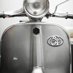 Puede incluir: Primer plano de la parte delantera de un scooter plateado. El scooter tiene un faro redondo, manillar cromado y carrocería plateada. Un hexágono negro y un emblema cromado con las letras "B" y "H" son visibles en la parte delantera.