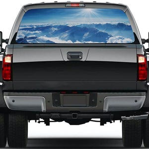 Puede incluir: Camioneta negra con una ventana trasera que muestra una vista panorámica de montañas y cielo. La imagen muestra un sol brillante sobre picos montañosos cubiertos de nieve y nubes blancas. La camioneta tiene ruedas traseras dobles y espejos laterales.