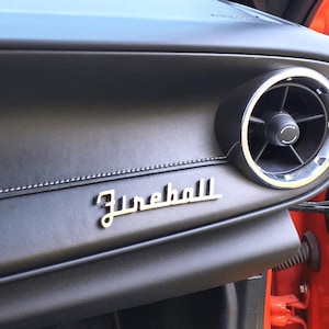 May include: Close-up of a black leather car dashboard with a silver "Fireball" emblem and a round air vent.