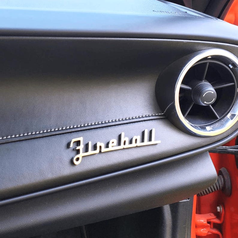 May include: Close-up of a black leather car dashboard with a silver "Fireball" emblem and a round air vent.