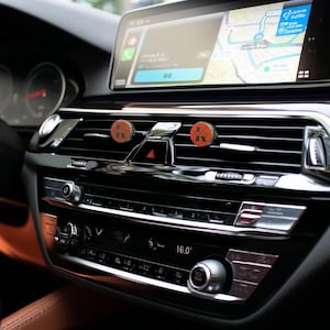 Könnte beinhalten: Autoinnenraum mit zwei orangefarbenen Lufterfrischern mit einem "K"-Design. Das Armaturenbrett umfasst einen Navigationsbildschirm, Lüftungsschlitze und Klimabedienelemente. Die Temperaturanzeige zeigt 16,0 Grad Celsius an.