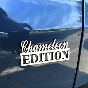 May include: A silver metal badge with the words "Chameleon Edition" in black and white. The badge is attached to a blue car.