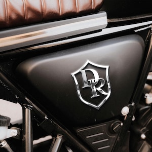 Op de afbeelding: Detail van een motorfiets zijpaneel met een zilveren "DR" embleem in een schildvorm. Het paneel is matzwart, het embleem glanzend. Een bruine leren zitting is zichtbaar op de achtergrond. Het frame is zwart.